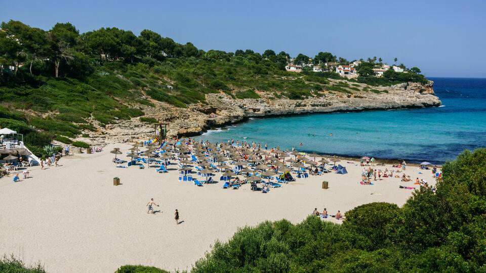 A crowded beach with clear turquoise water, surrounded by rocky cliffs and lush greenery.