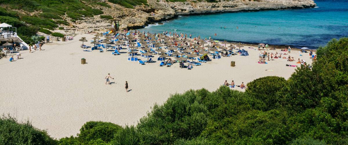 A crowded beach with clear turquoise water, surrounded by rocky cliffs and lush greenery.
