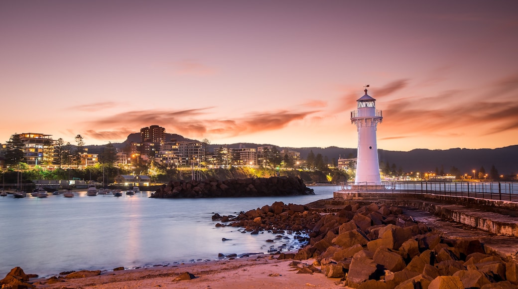 A coastal town at dusk with a prominent white lighthouse, rocky shoreline, and illuminated buildings.
