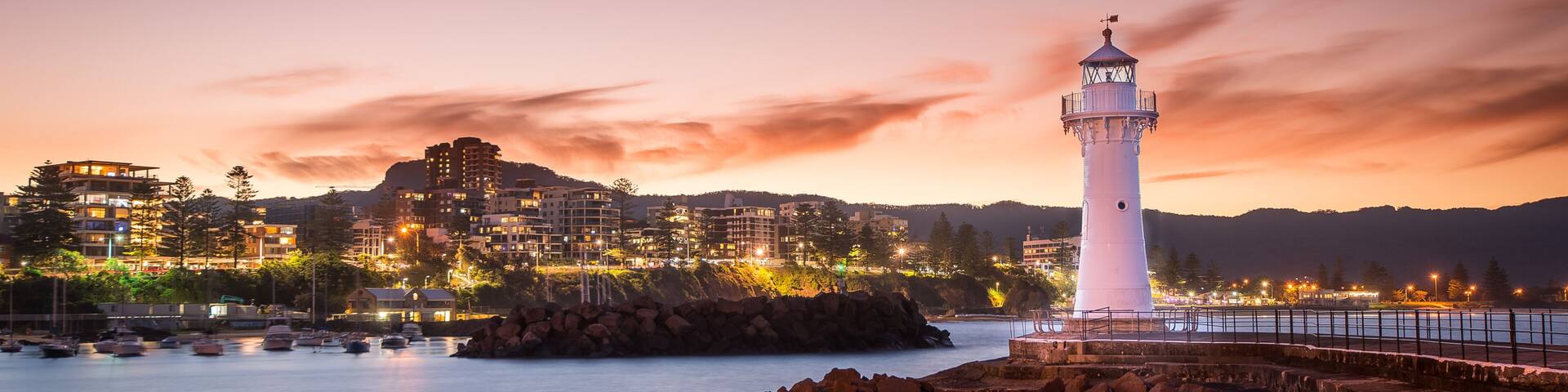 A coastal town at dusk with a prominent white lighthouse, rocky shoreline, and illuminated buildings.
