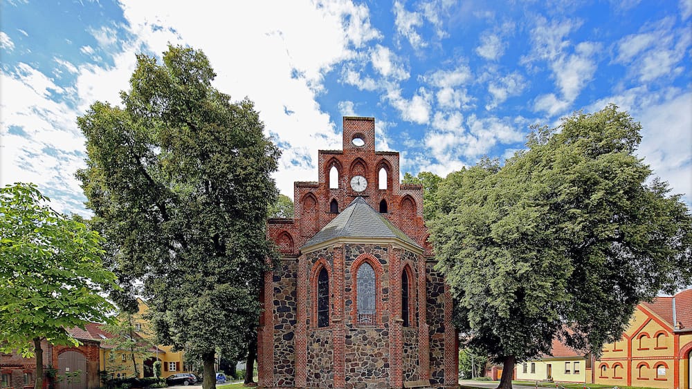 Eine historische Backsteinkirche mit einem Glockenturm, umgeben von großen Bäumen und blauem Himmel.