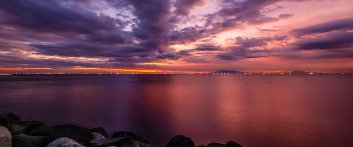 Un coucher de soleil sur une mer calme, avec une côte rocheuse et les lumières d'une ville lointaine.