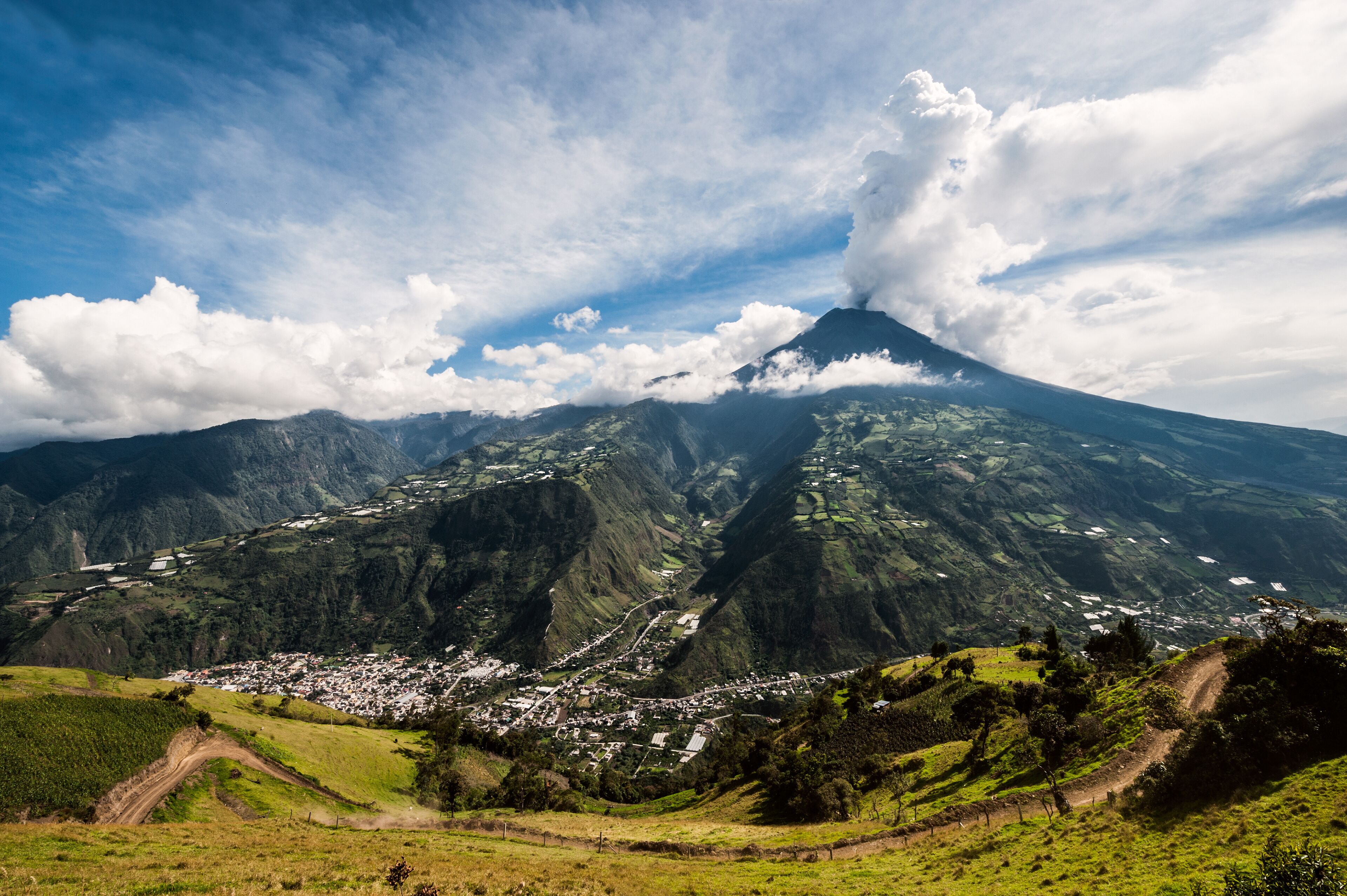 A mountain with a village at its base, surrounded by lush green hills and a partly cloudy sky.