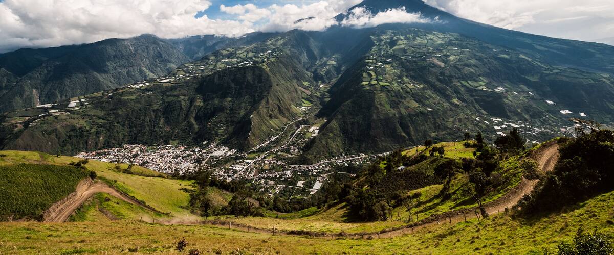 A mountain with a village at its base, surrounded by lush green hills and a partly cloudy sky.