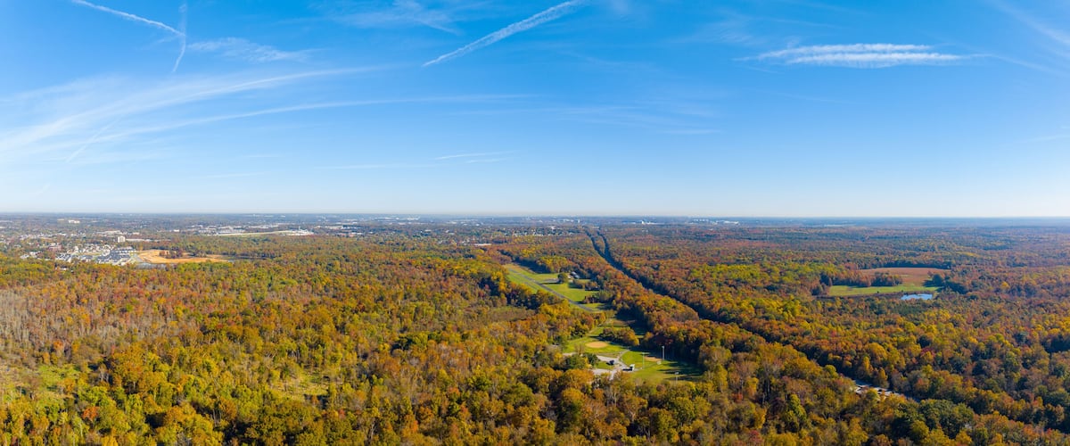 A panoramic view of a forested valley with a winding road, a small town in the distance, and a clear blue sky.