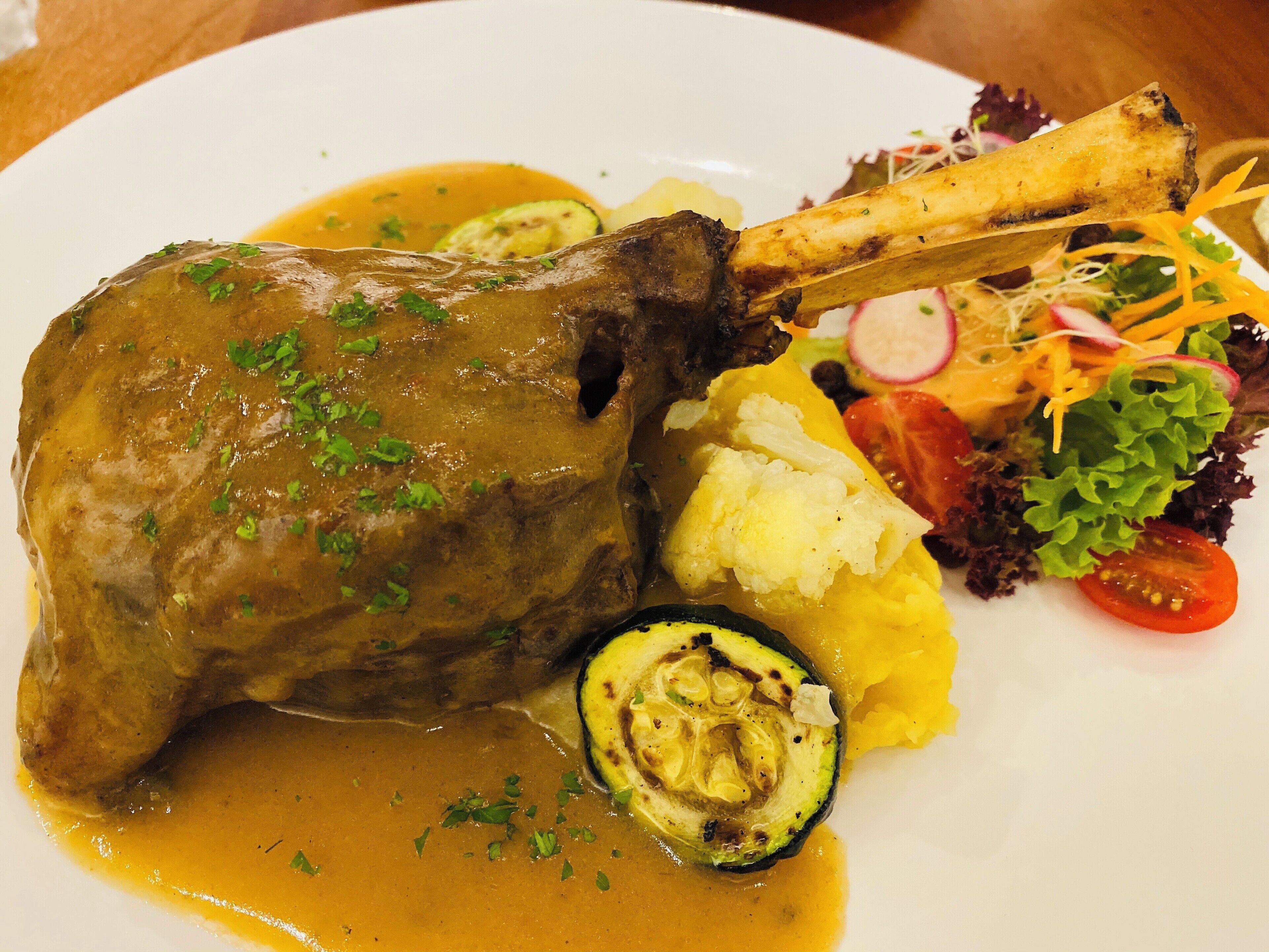A plate with a lamb shank, mashed potatoes, zucchini, and a side salad.