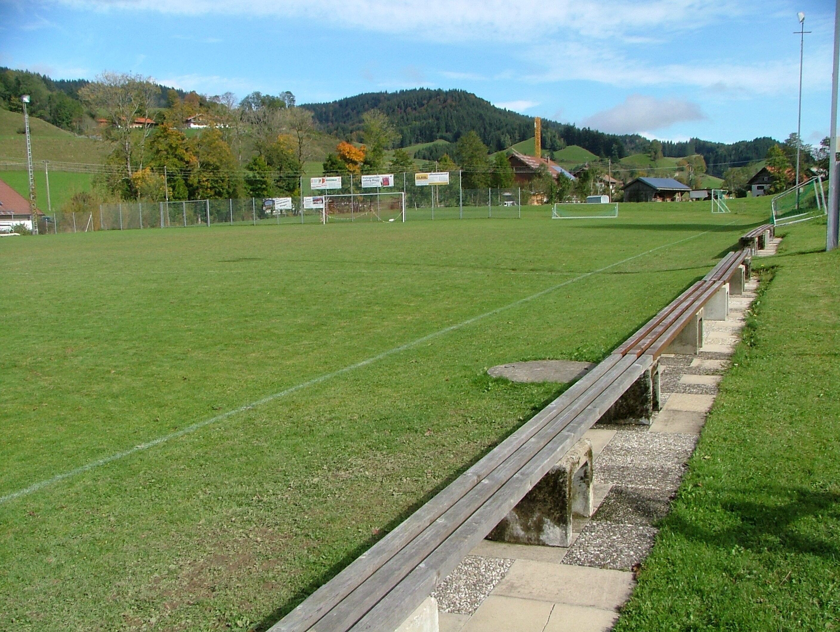 A grassy sports field with a wooden bench, surrounded by a fence and houses in the background.