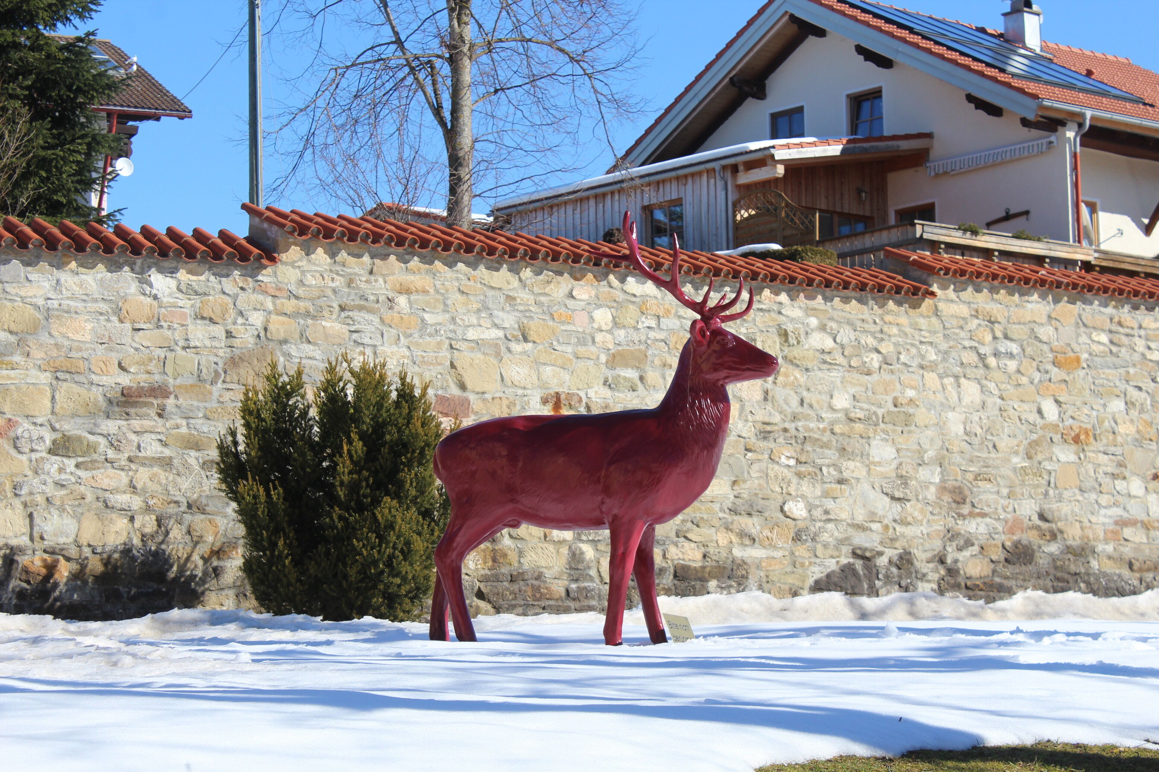 A red deer statue in a snowy area with a stone wall and a house in the background.