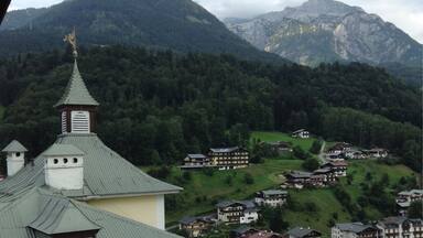 A mountain village with traditional houses, a church with a green roof, and a forested mountain backdrop.