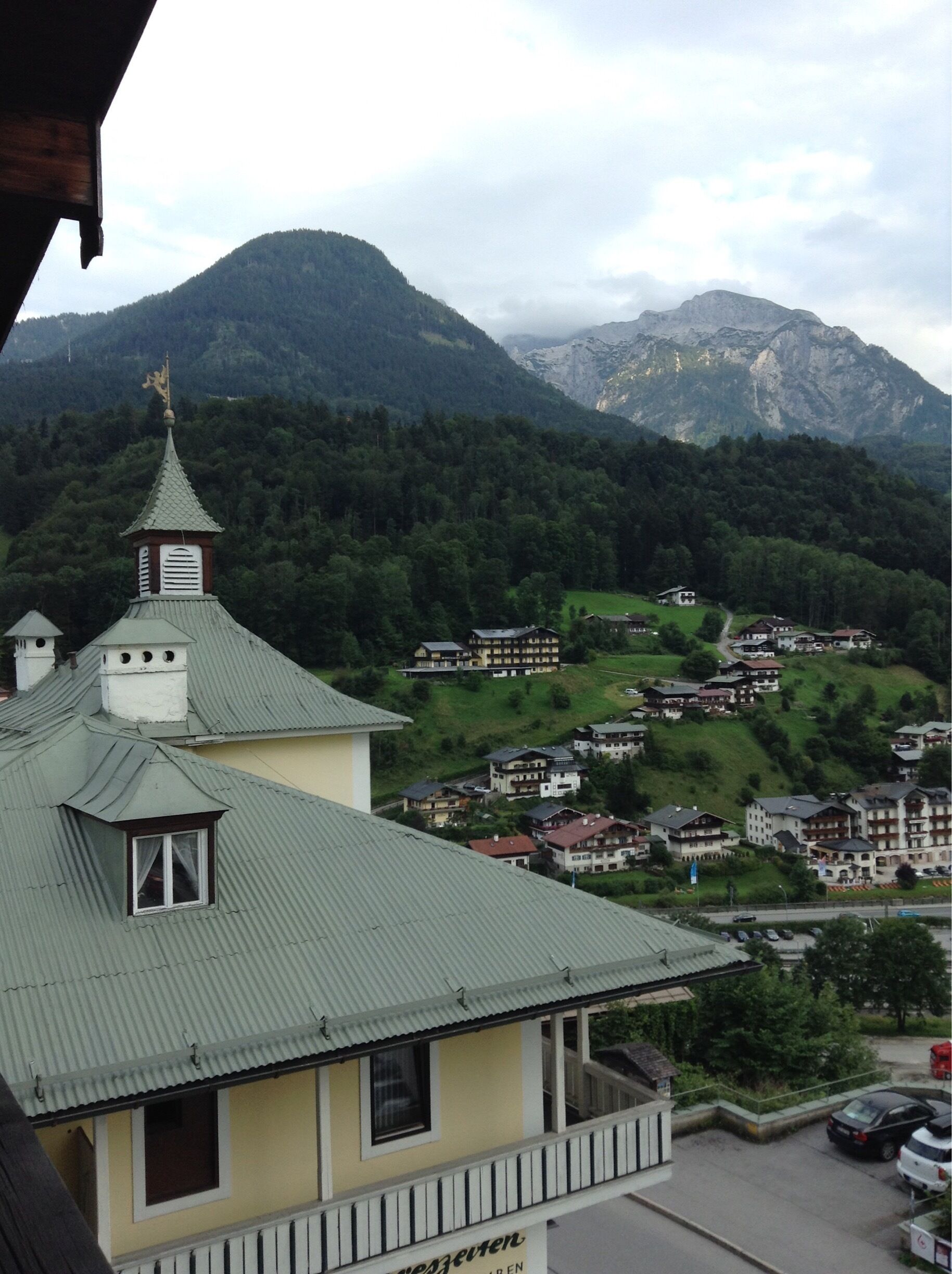 A mountain village with traditional houses, a church with a green roof, and a forested mountain backdrop.