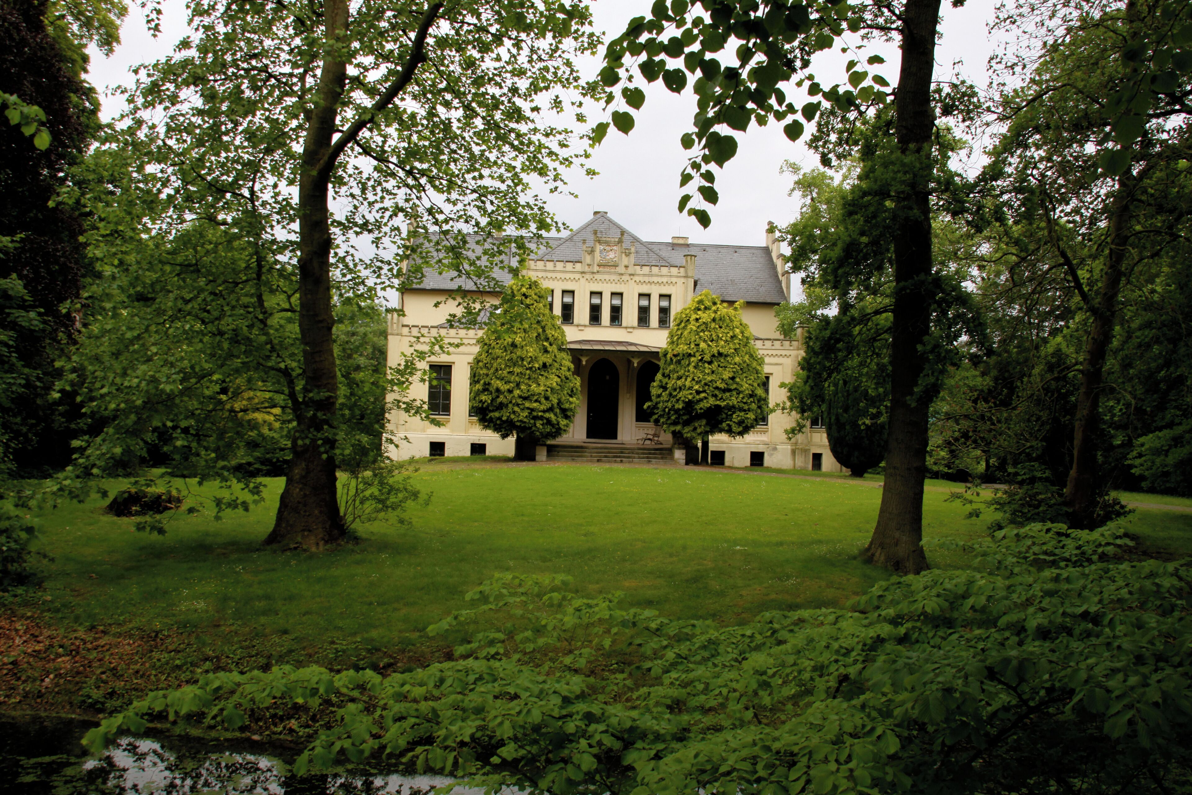 A stately building with a symmetrical facade, surrounded by lush greenery and two prominent trees framing the entrance.