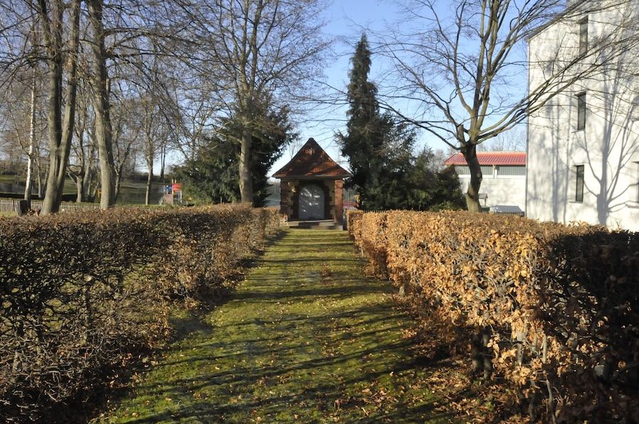 Ein Steinweg führt zu einem kleinen, aus Ziegeln gebauten Gebäude, das von Bäumen und Hecken umgeben ist.