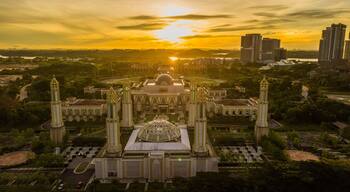 A grand building with a central dome and four tall towers, surrounded by greenery and modern high-rises in the background.