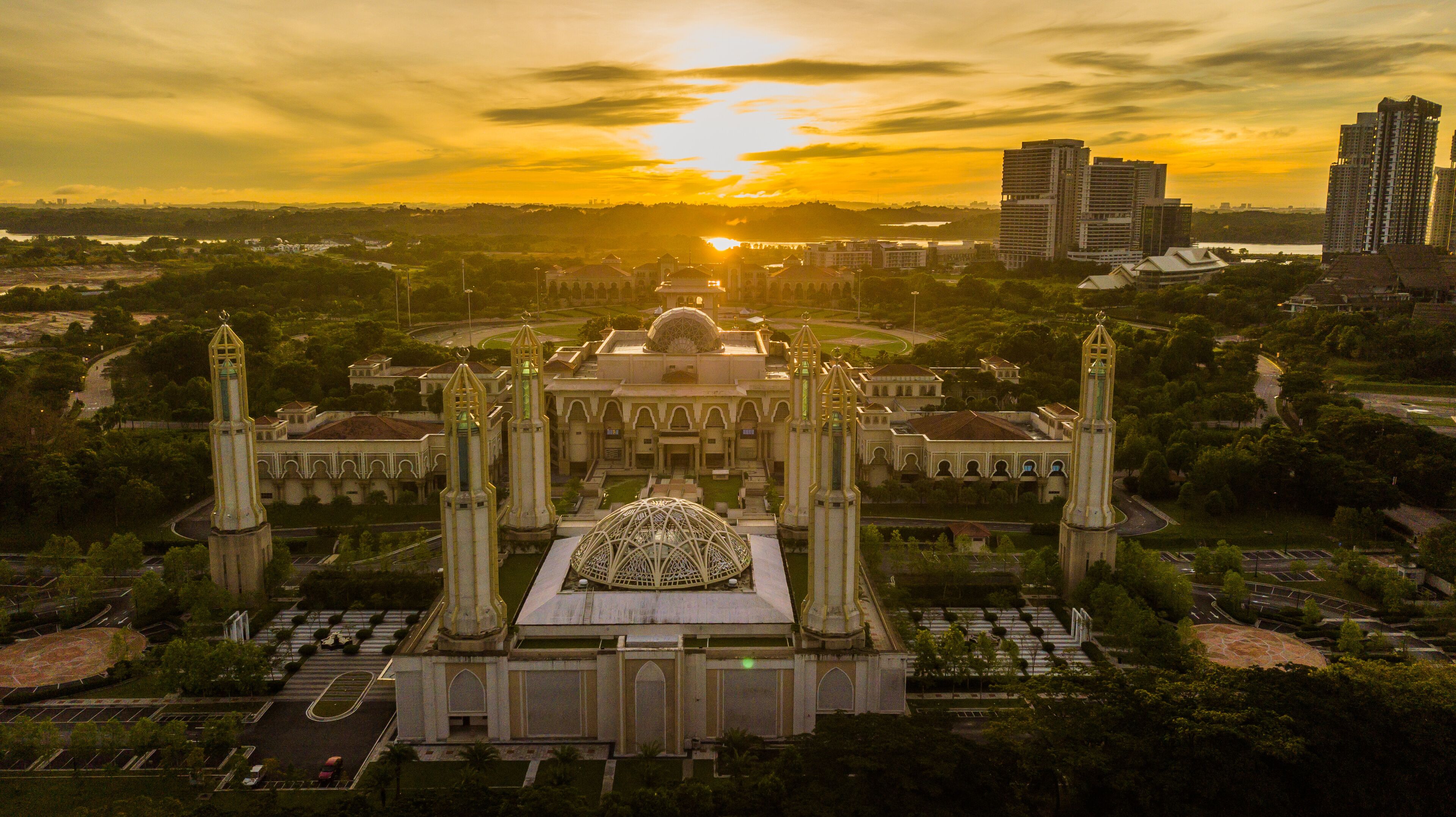 A grand building with a central dome and four tall towers, surrounded by greenery and modern high-rises in the background.