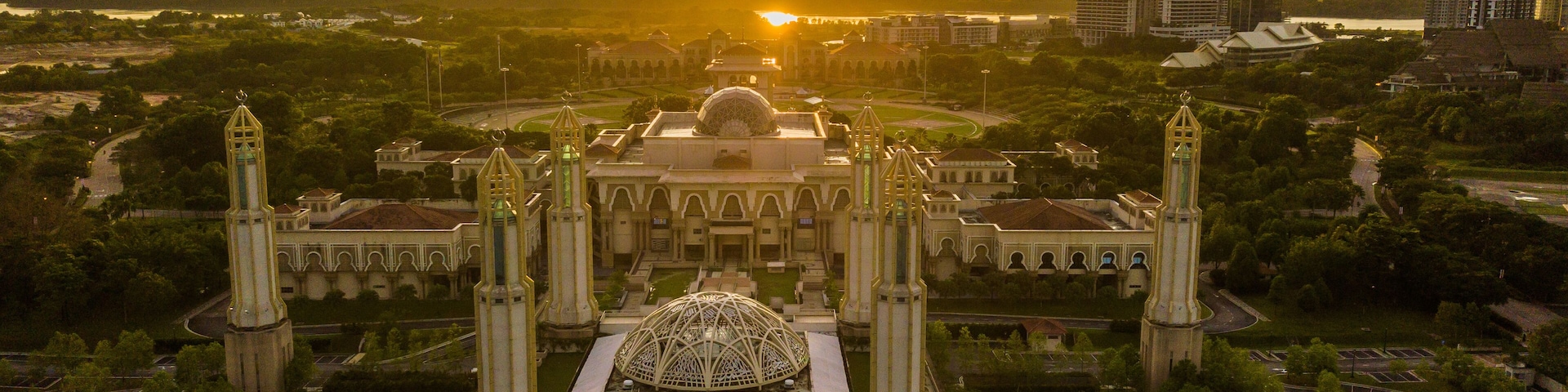 A grand building with a central dome and four tall towers, surrounded by greenery and modern high-rises in the background.