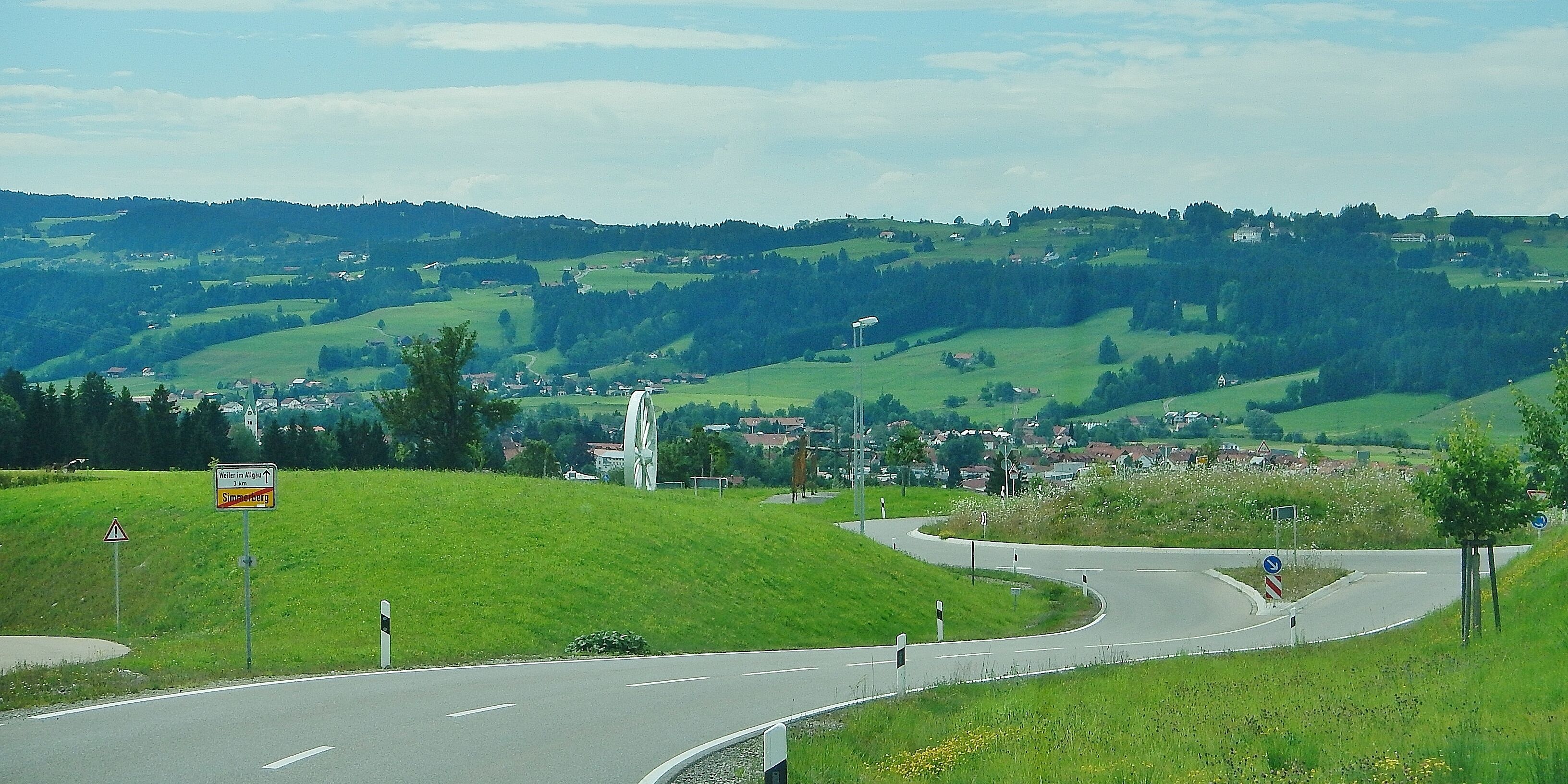 A winding road through a hilly landscape with a large wind turbine and a village in the distance.