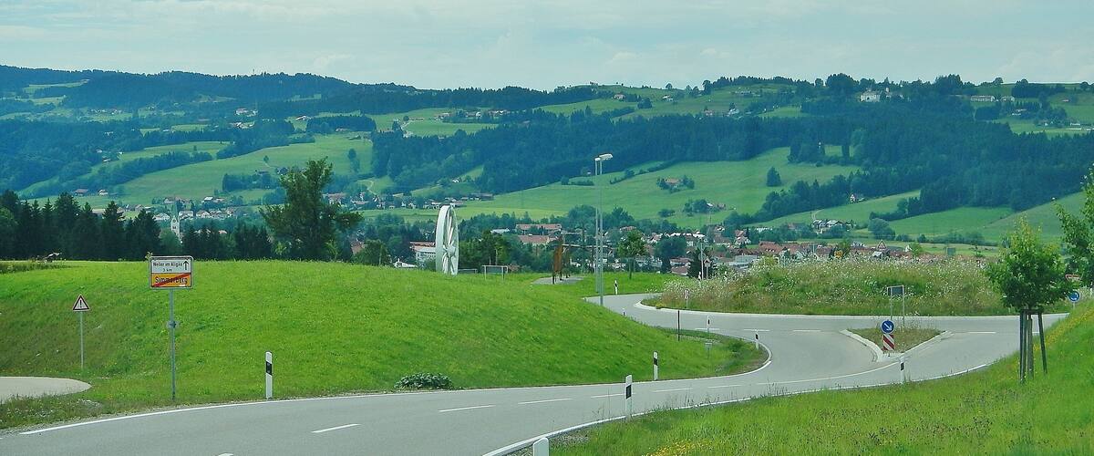 A winding road through a hilly landscape with a large wind turbine and a village in the distance.