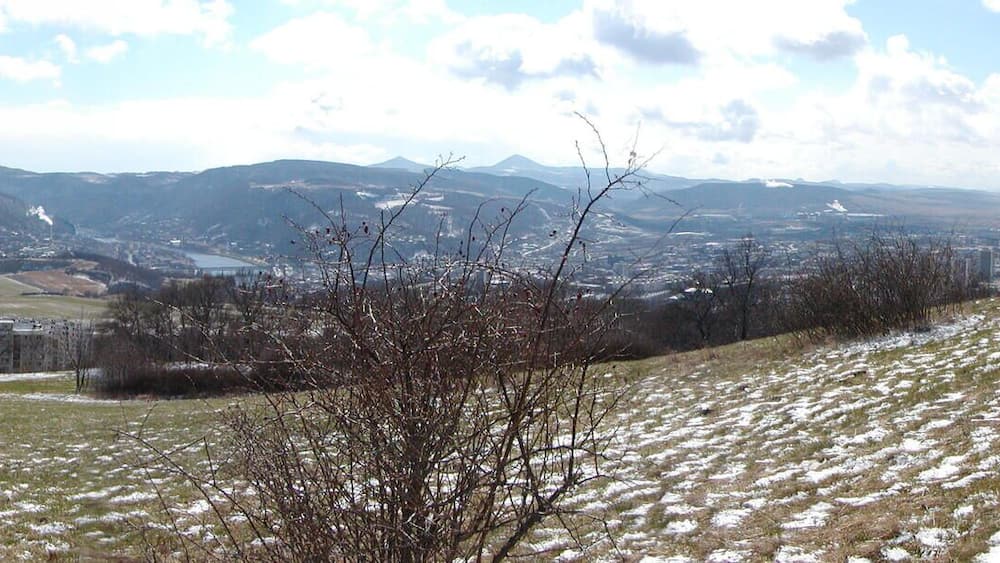 A hilly landscape with a town in the valley, snow patches, and distant mountains.