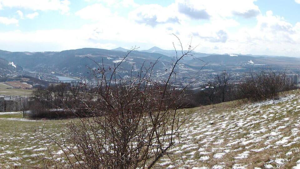 A hilly landscape with a town in the valley, snow patches, and distant mountains.