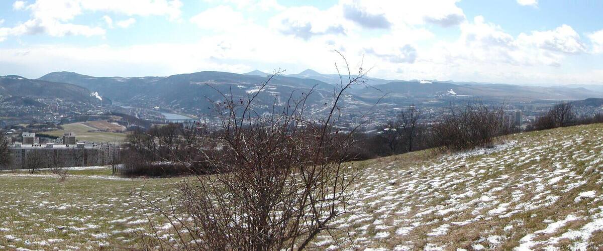 A hilly landscape with a town in the valley, snow patches, and distant mountains.