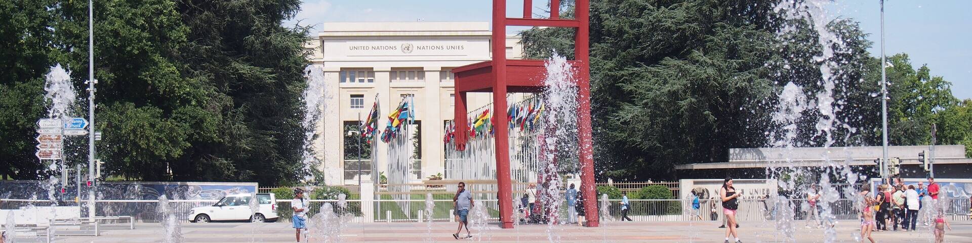 A public square with a large red sculpture, fountains, and a building with flags.
