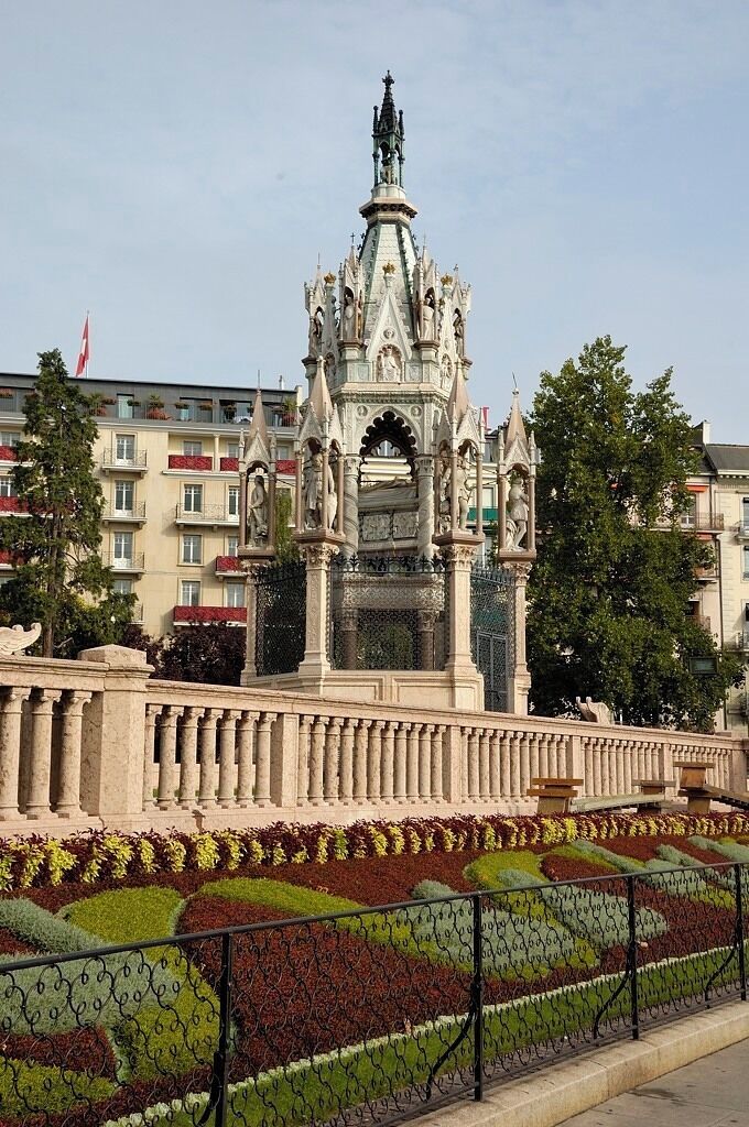 A Gothic-style monument with intricate carvings and a statue on top, surrounded by a landscaped garden with geometric patterns.