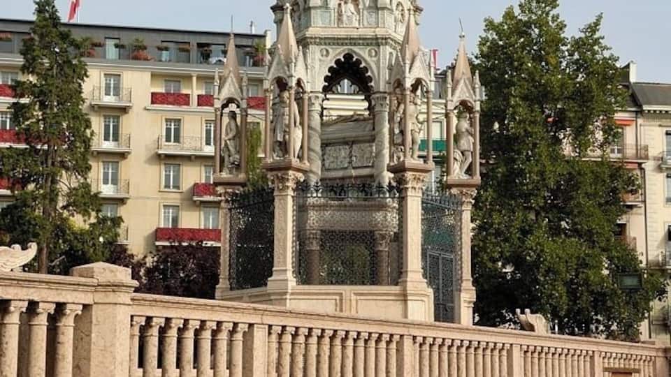 A Gothic-style monument with intricate carvings and a statue on top, surrounded by a landscaped garden with geometric patterns.