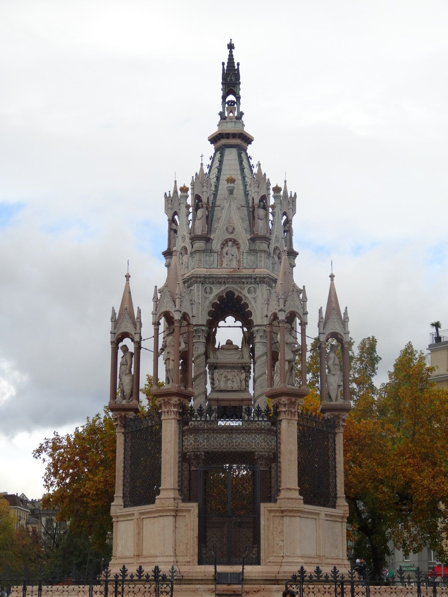 Um monumento em estilo gótico com esculturas e entalhes elaborados, rodeado por árvores e uma cerca.