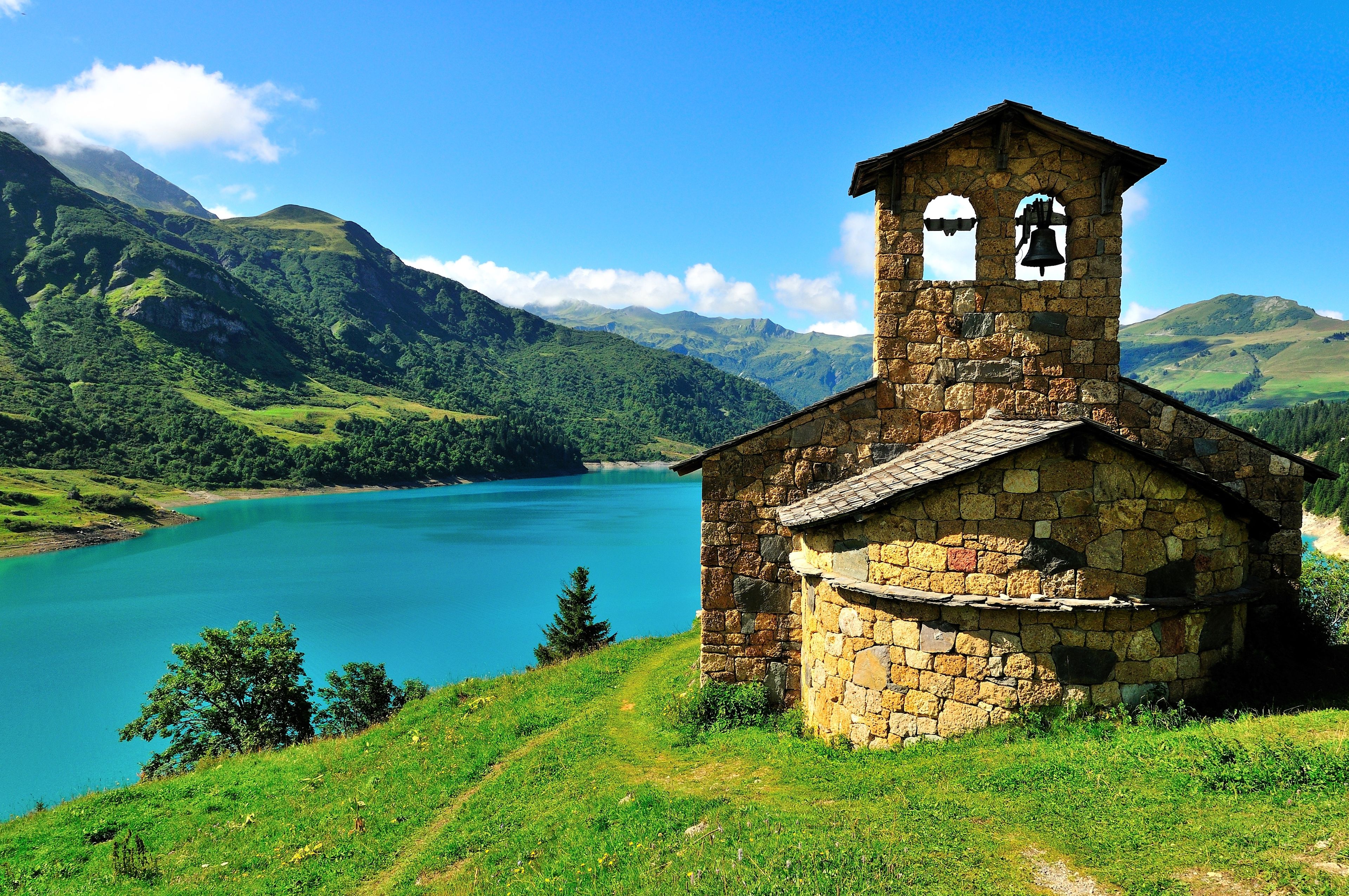 A stone church with a bell tower, surrounded by lush green hills and a clear blue lake.