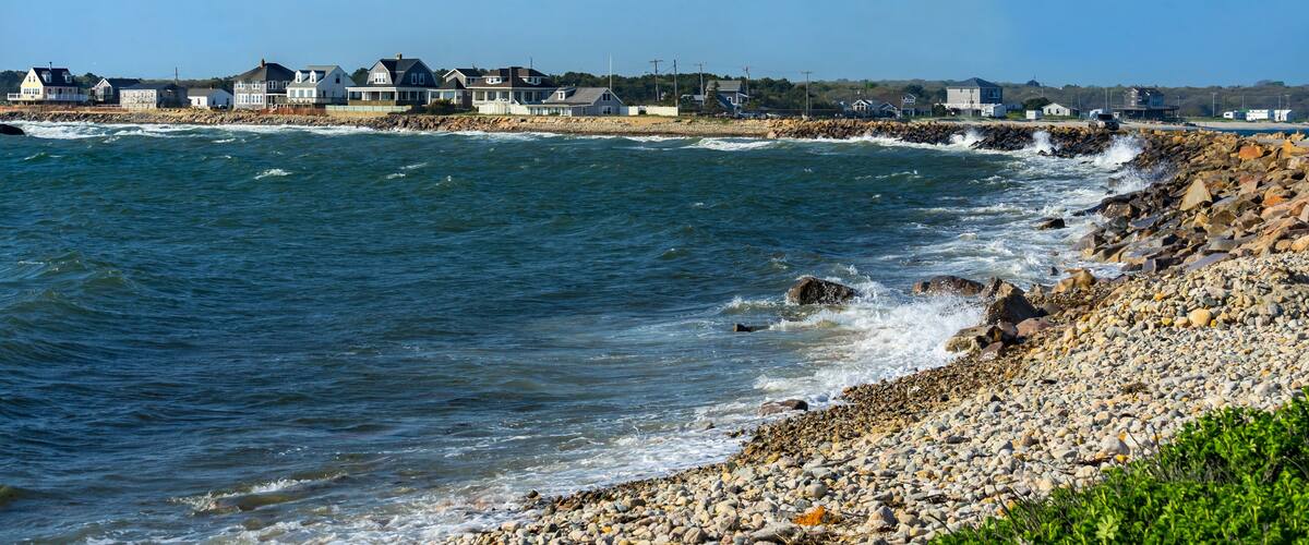 A rocky shoreline with waves crashing onto the beach, houses along the coast, and a clear blue sky.