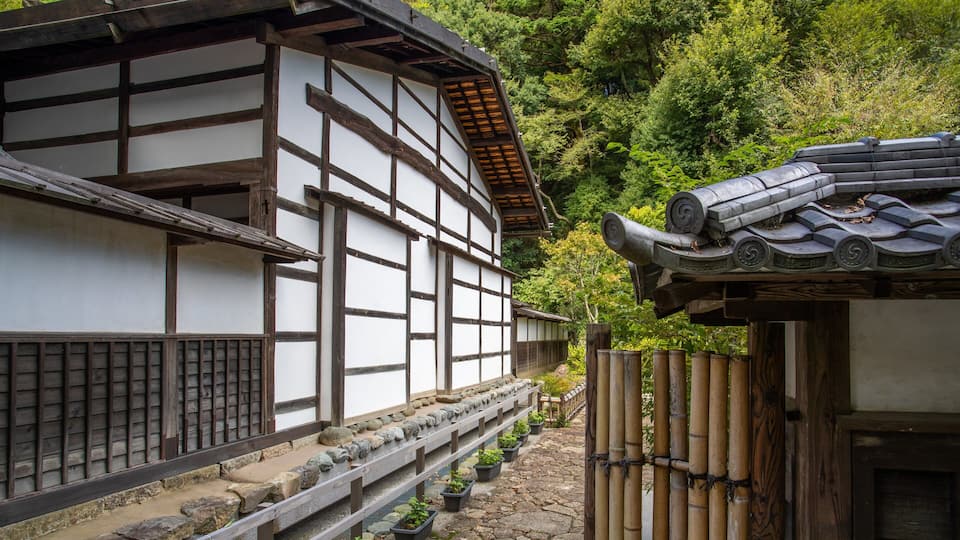 Edificio japonés tradicional con paredes blancas y vigas de madera oscura, rodeado de vegetación exuberante y escalones de piedra que conducen a una cerca de madera.