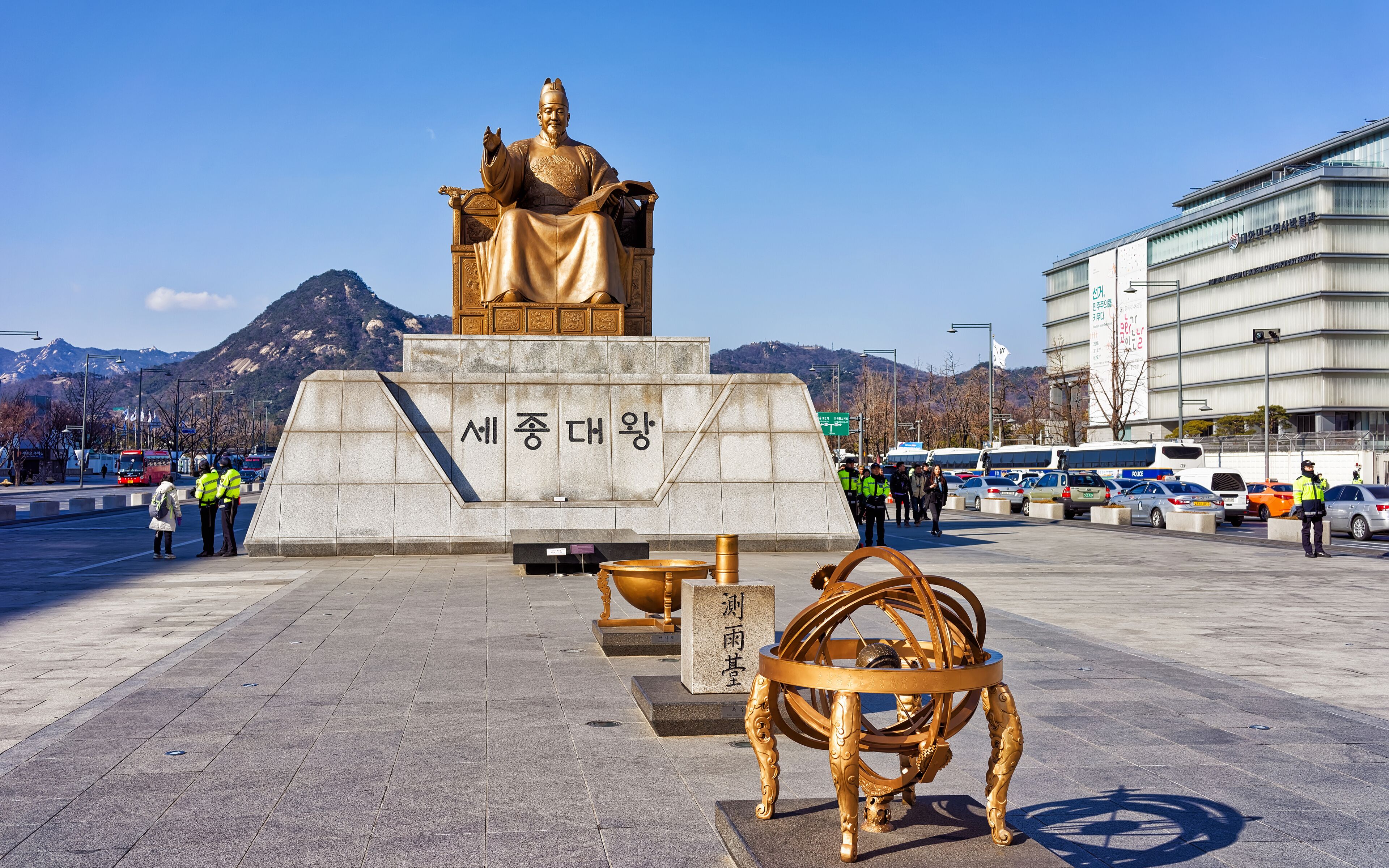 A statue of a seated figure with inscriptions, a golden astronomical instrument, and a modern building in the background.