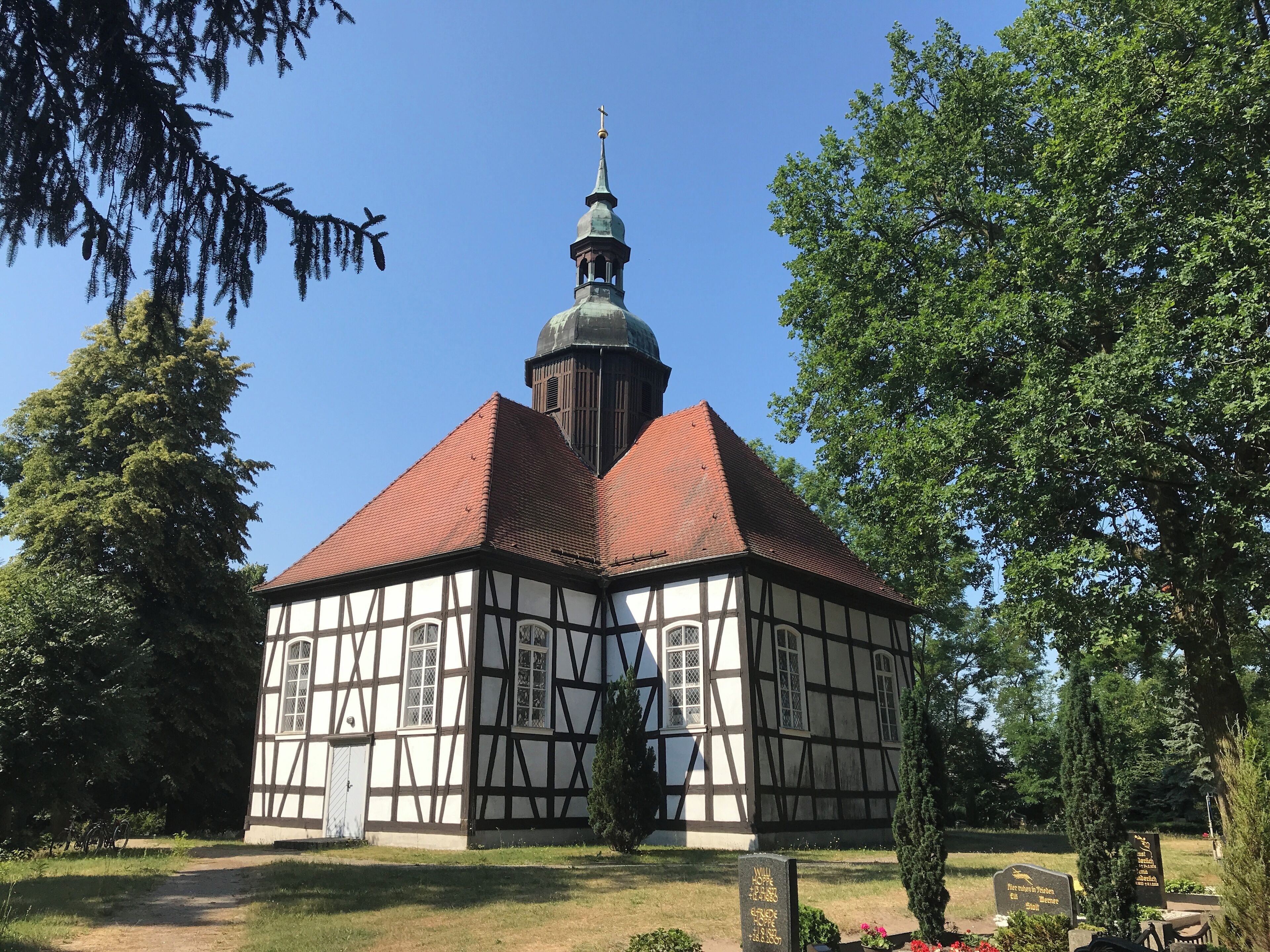 A half-timbered church with a red roof and a dome, surrounded by trees and a clear blue sky.