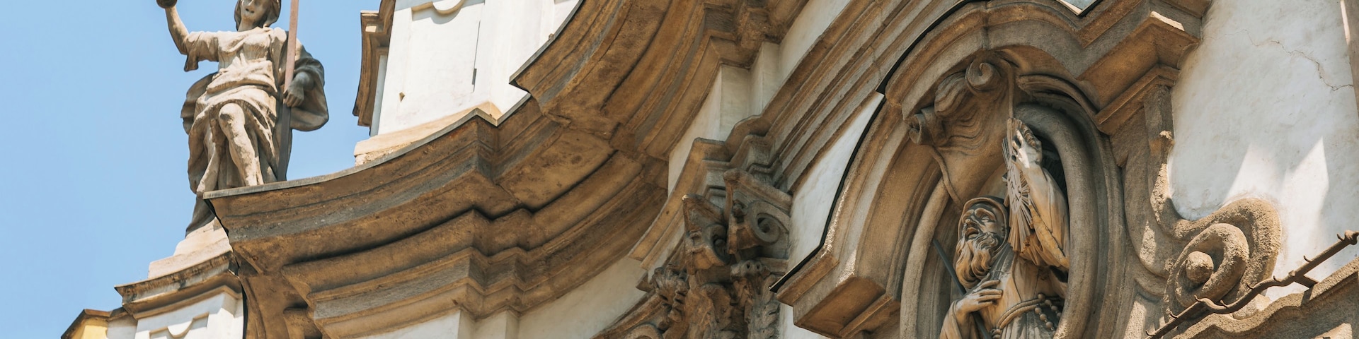 Eine detaillierte Steinfassade mit einer Statue, die eine Figur zeigt, die ein Kreuz und ein Buch hält, sowie eine weitere Figur, die ein Saiteninstrument spielt.