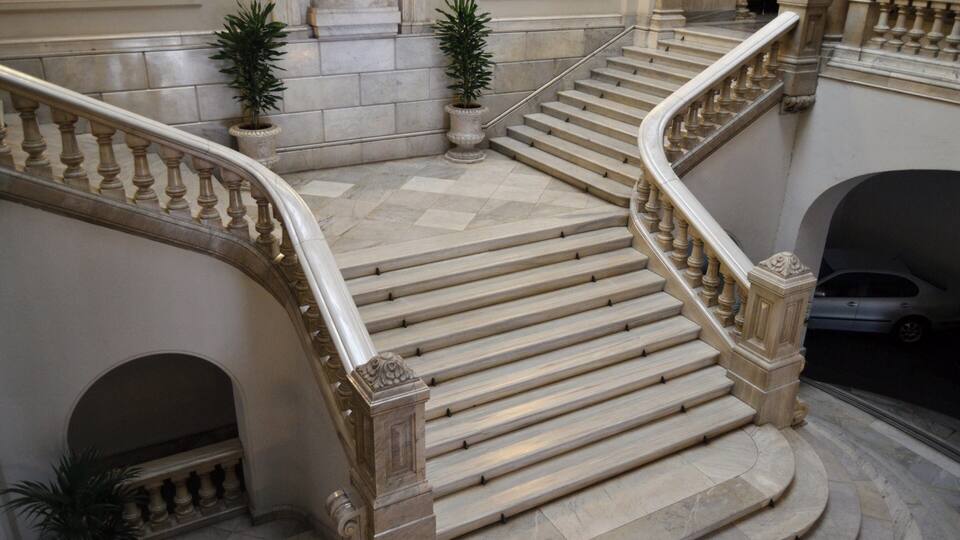 Una imponente escalera de mármol con barandillas ornamentales, flanqueada por macetas con plantas.