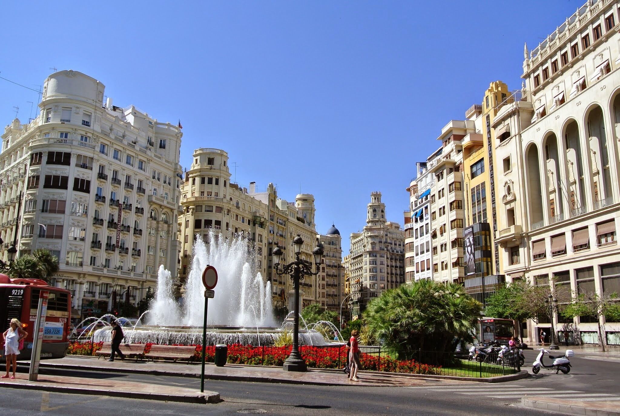A city square with a large fountain, historic buildings, and a clear blue sky.