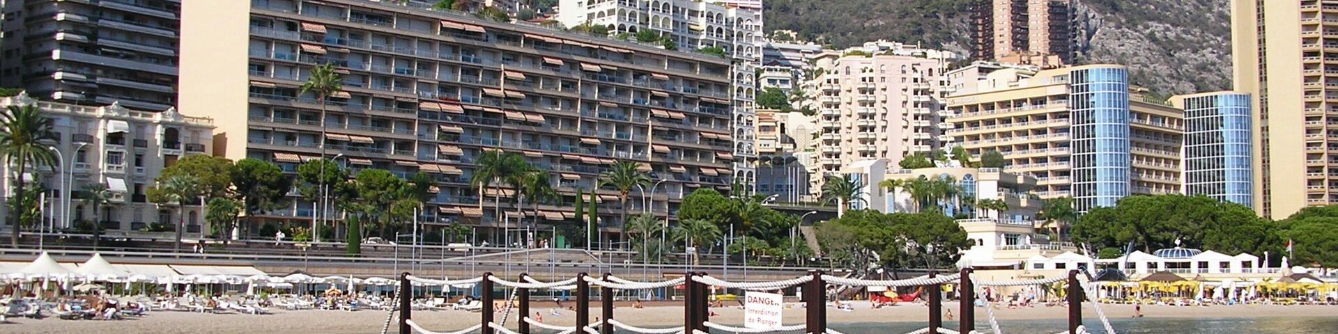 Un paisaje urbano costero con un hotel destacado, montañas rocosas al fondo y un muelle con barandilla de seguridad.