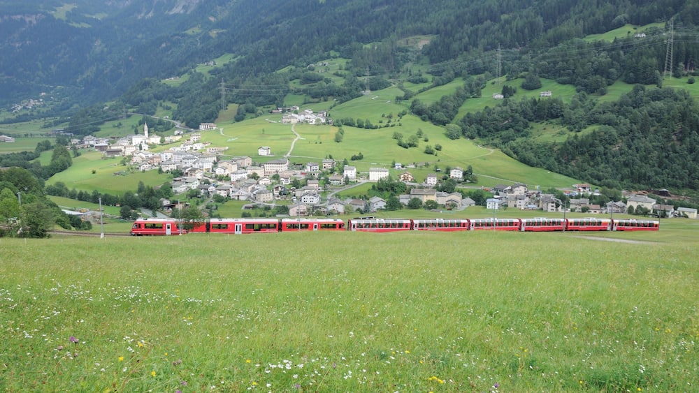 Un train rouge circule sur une voie ferrée qui traverse une vallée verdoyante, avec une ville en arrière-plan.