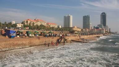 A crowded beach with waves crashing onto the shore, a sandy promenade, and tall buildings in the background.