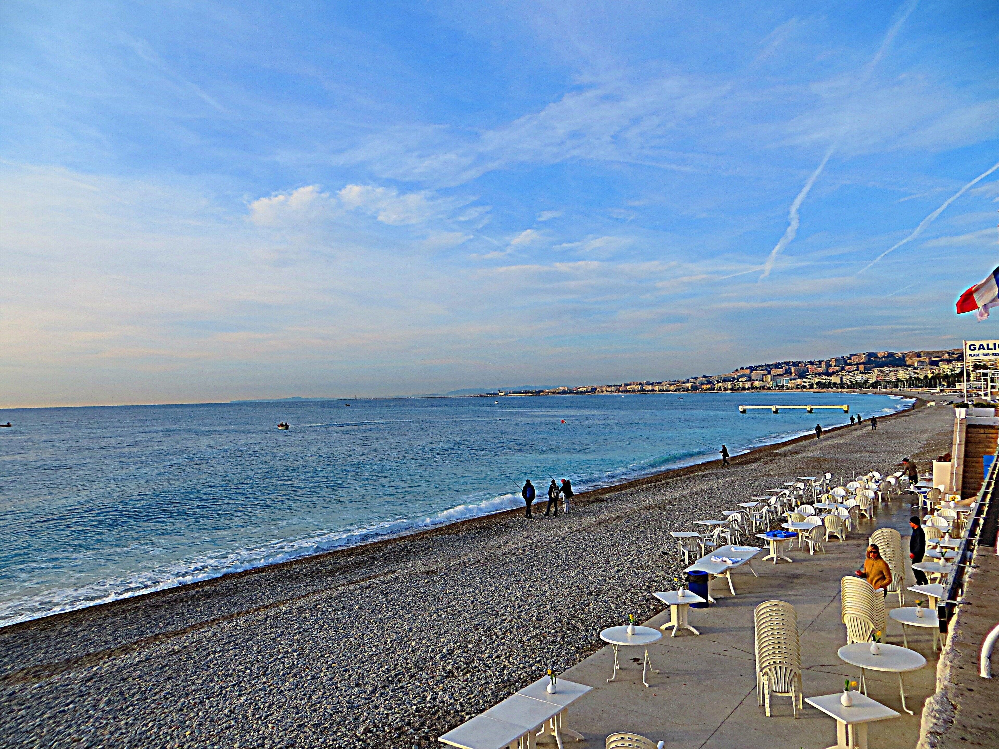 A pebbled beach with tables and chairs, a clear sky, and a distant cityscape.