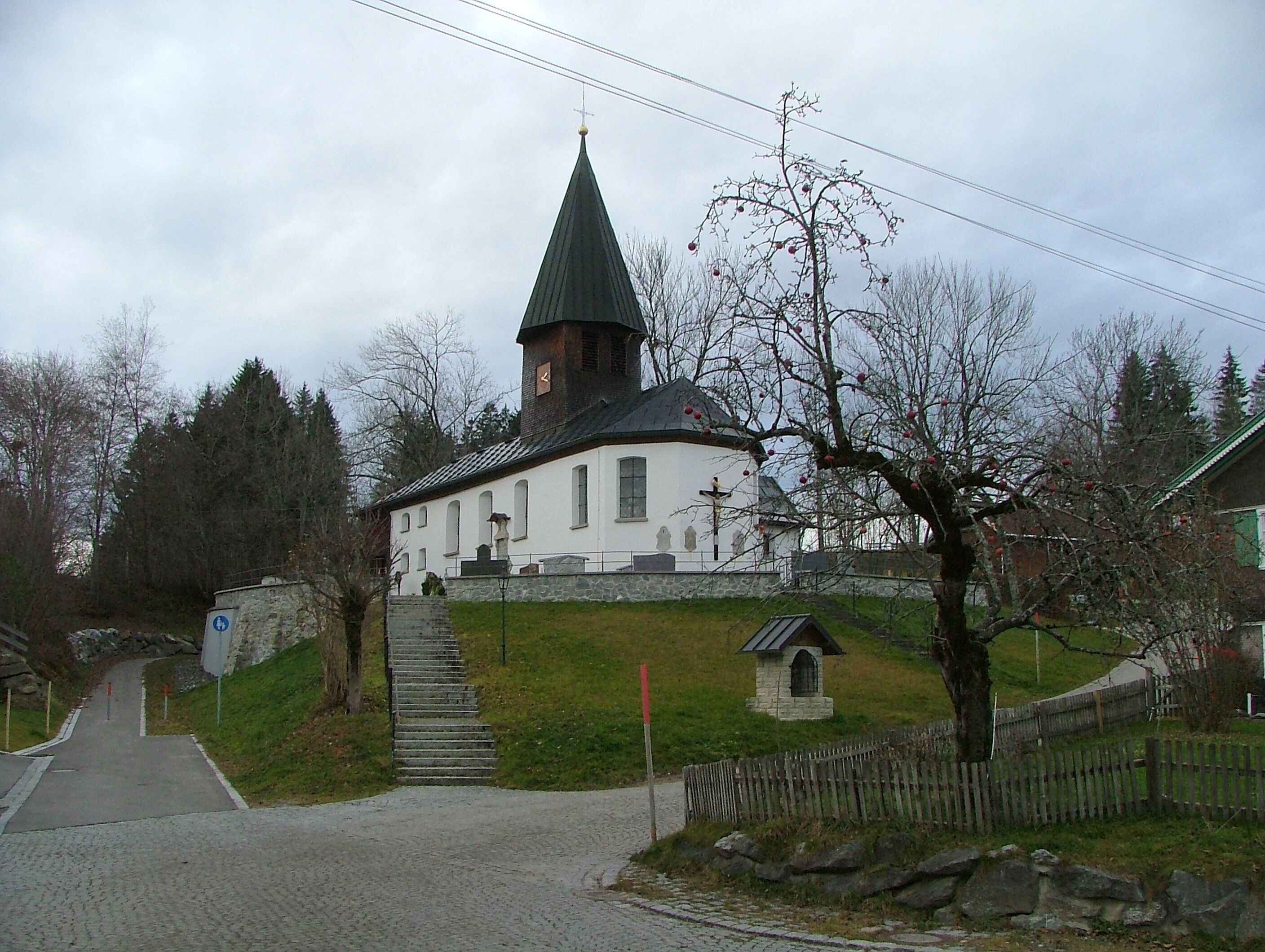A church with a steeply pitched roof and a bell tower, surrounded by trees and a stone wall.