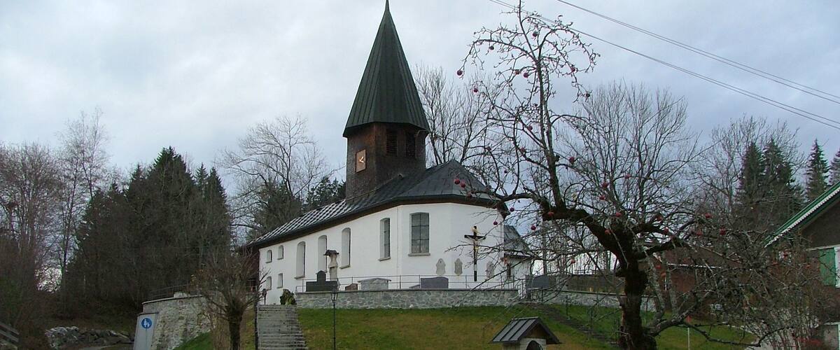 A church with a steeply pitched roof and a bell tower, surrounded by trees and a stone wall.
