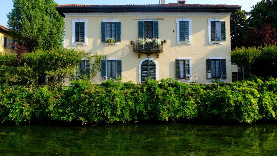 Ein großes, cremefarbenes Gebäude mit grünen Fensterläden, umgeben von üppigem Grün und einer ruhigen Wasserfläche.