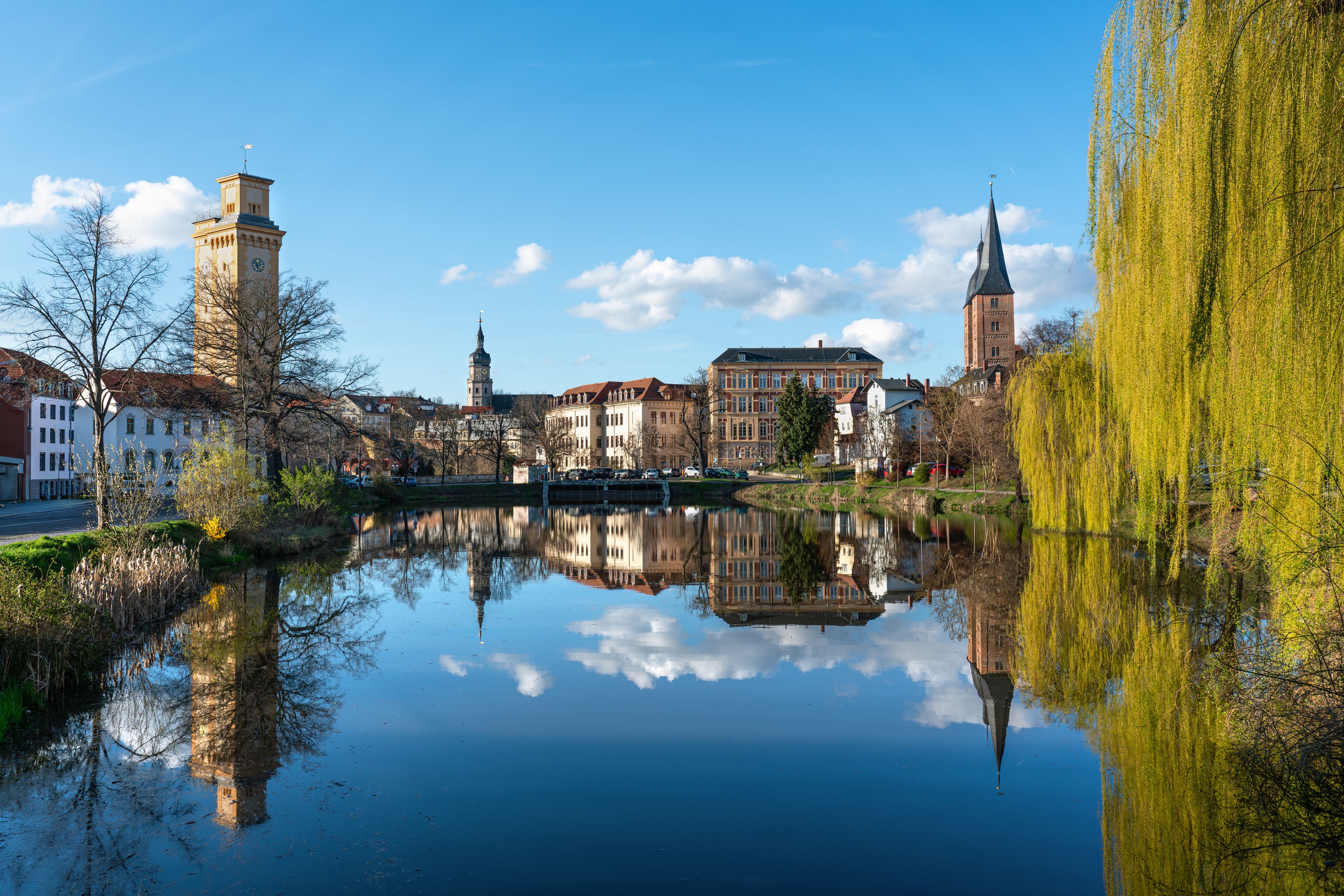 Une rivière paisible où se reflètent des bâtiments, un beffroi et le clocher d’une église.