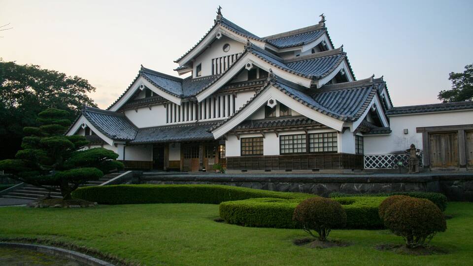 A traditional Japanese castle with multiple tiers, wooden balconies, and a well-maintained garden with trimmed bushes and a pond.