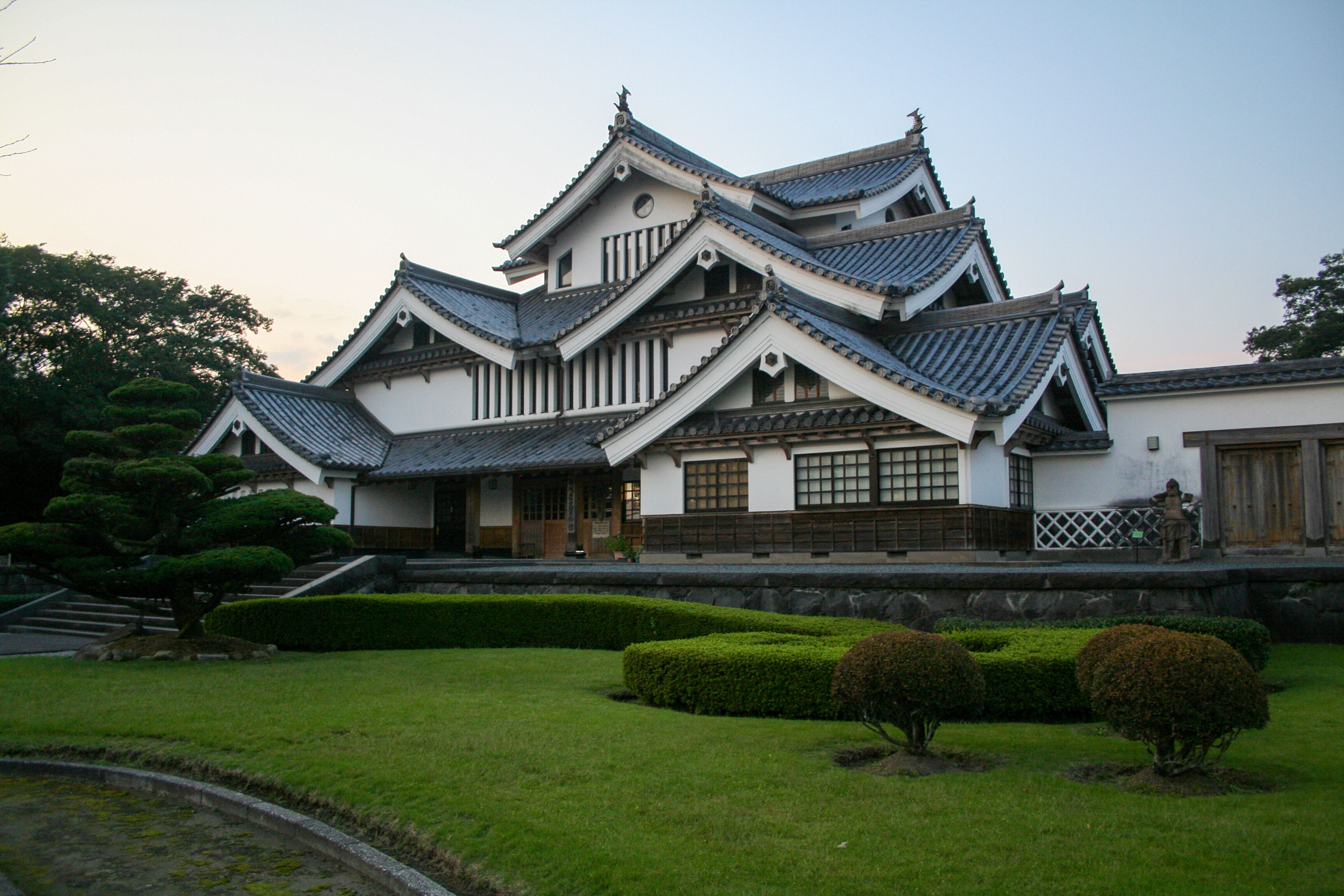 A traditional Japanese castle with multiple tiers, wooden balconies, and a well-maintained garden with trimmed bushes and a pond.