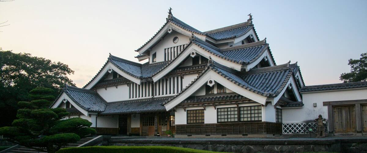 A traditional Japanese castle with multiple tiers, wooden balconies, and a well-maintained garden with trimmed bushes and a pond.
