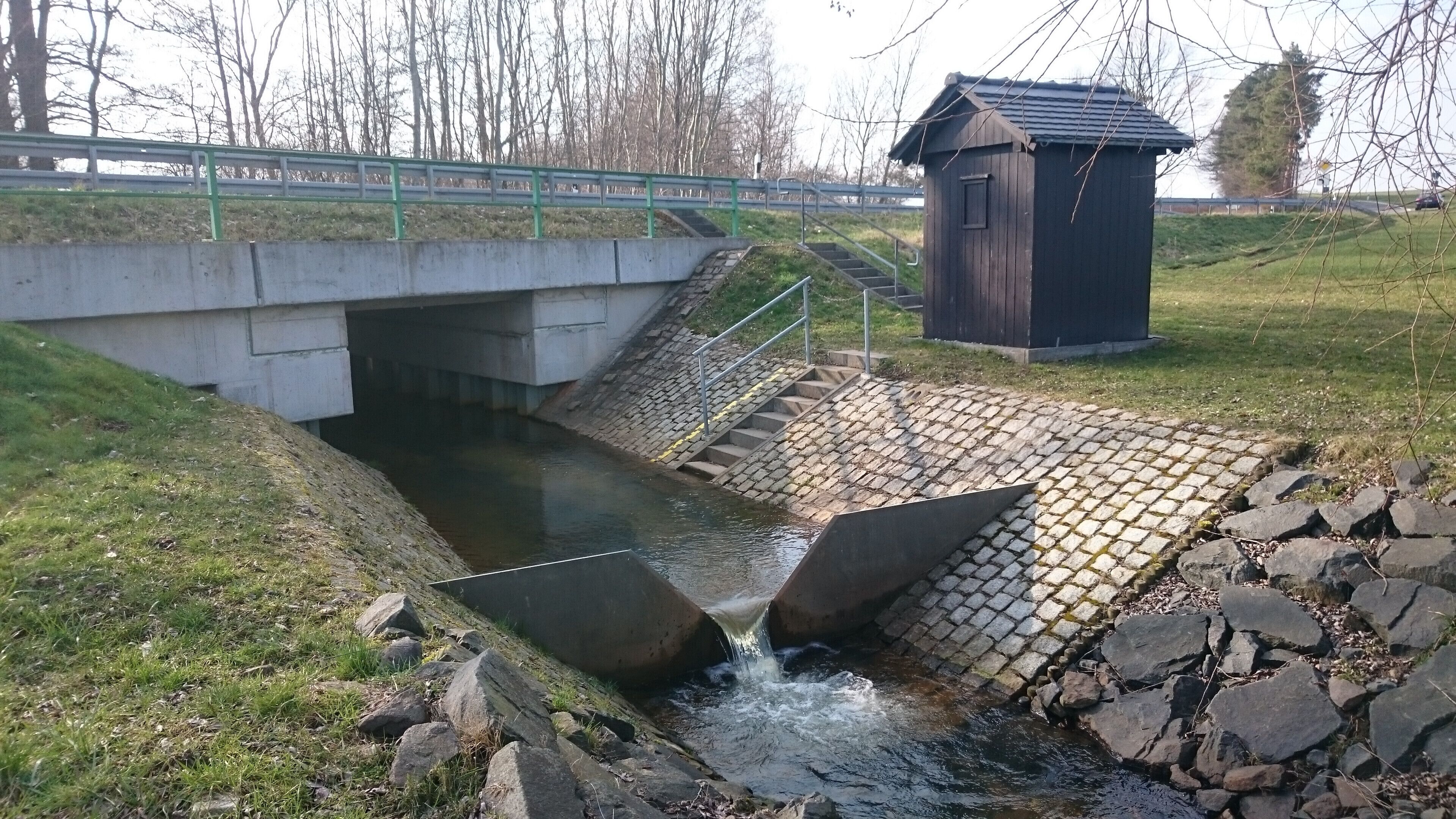 A concrete bridge over a small stream with a wooden structure nearby.