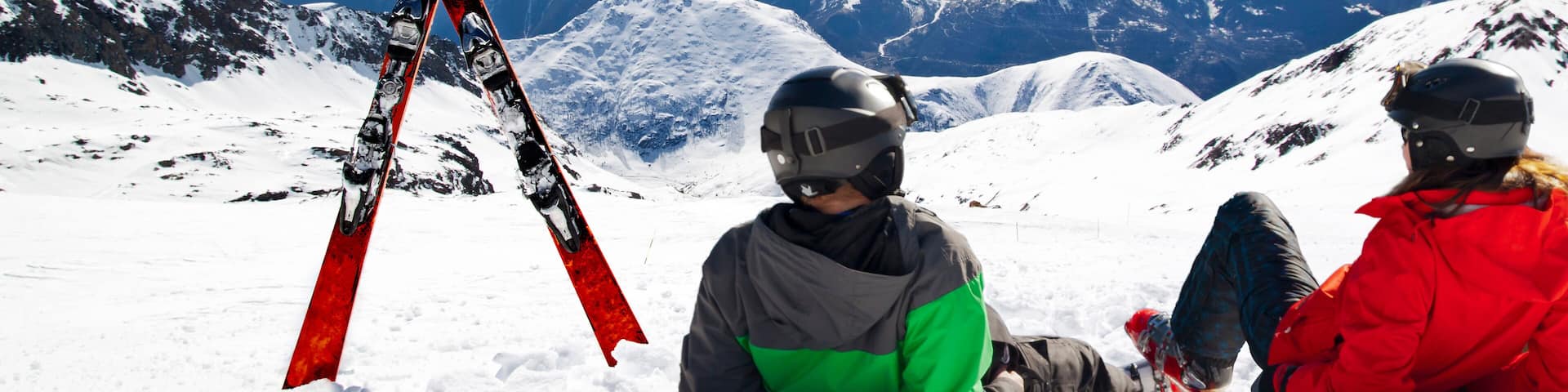 Two people sitting on a snowy mountain with a pair of skis leaning against them, and a mountain range in the background.