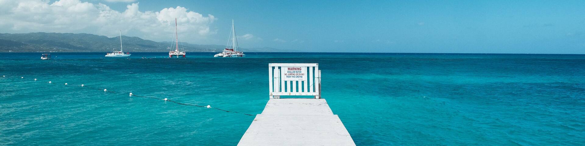 A pier leading into turquoise waters with boats in the distance and a warning sign at the end.
